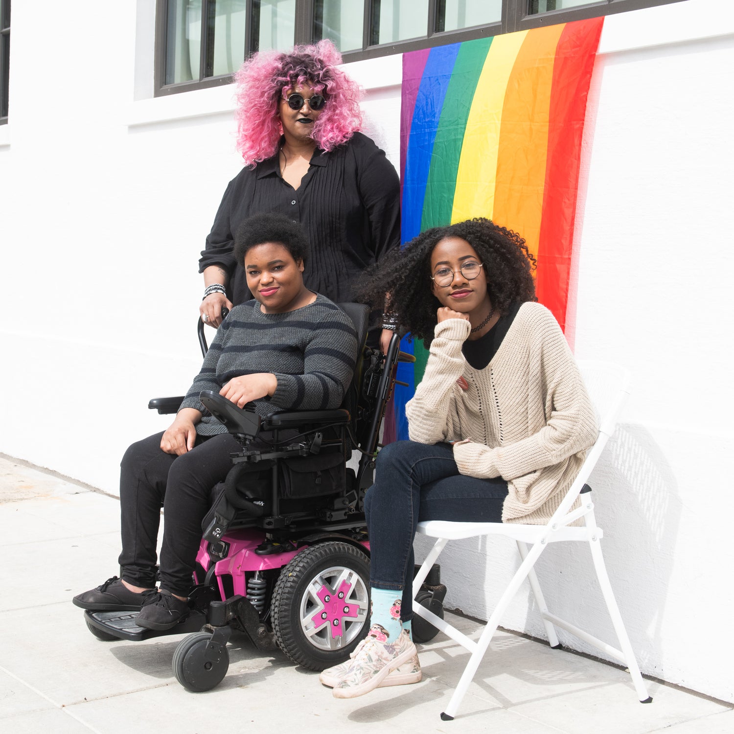 Three Black and disabled folx (a non-binary person holding a cane, a non-binary person sitting in a power wheelchair, and a femme sitting in a chair) casually smile at the camera while a rainbow pride flag drapes on the wall behind them.